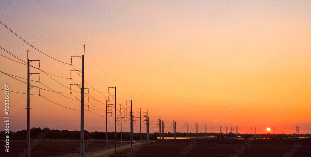 Silhouette two rows of many electric poles with cable lines along curve street in countryside area against orange sunset sky background, panoramic view