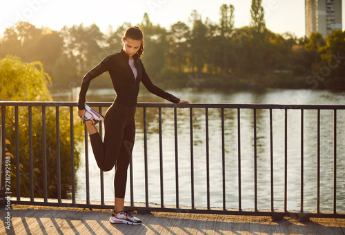 Woman with a slim sporty figure doing warming up exercises during an outdoor fitness workout in summer. Beautiful young girl in a sports suit doing leg exercises while standing by the bridge railing