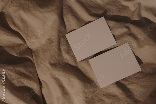 Blank paper card sheets with copy space on wrinkled bed blanket cloth with aesthetic warm sunlight shadows. Flat lay, top view