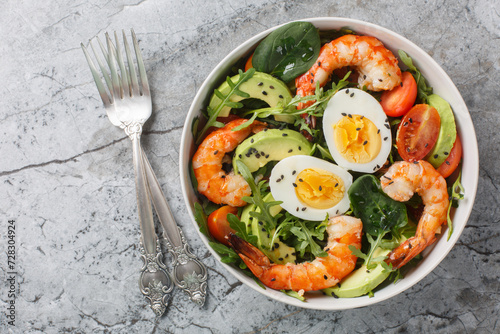 Delicious serving salad with shrimp, avocado, arugula, spinach, tomatoes, sesame seeds and boiled eggs close-up on a bowl on the table. Horizontal top view from above