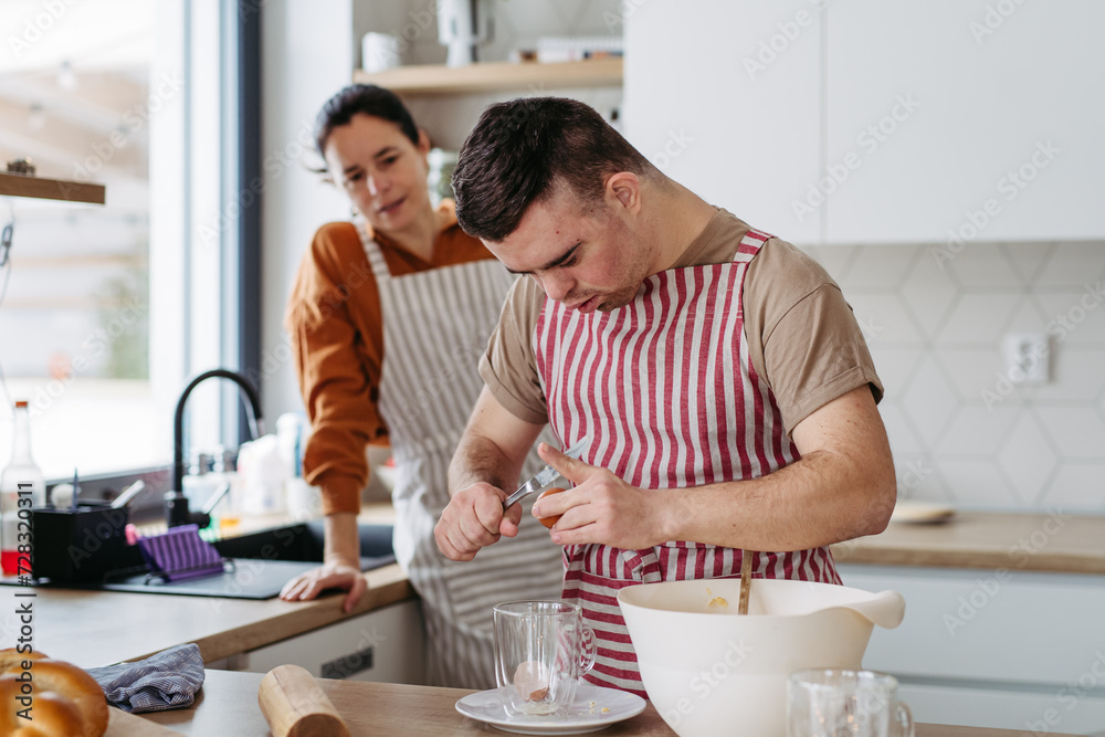 Young man with Down syndrome preparing breakfast with his mother at home, cracking eggs. Morning routine for man with Down syndrome. Young man with Down syndrome preparing breakfast with his mother at home, cracking eggs. Morning routine for man with Down syndrome.