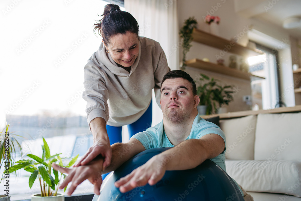 Young man with Down syndrome exercising at home with his mother on fitness ball. Workout routine for disabled man. Young man with Down syndrome exercising at home with his mother on fitness ball. Workout routine for disabled man.