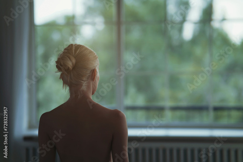 Naked young woman with blond hair looking through window photo