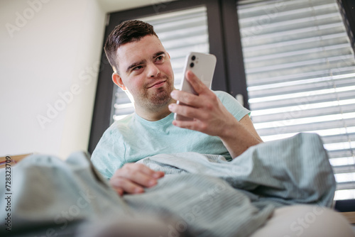 Young man with down syndrome lying in bed, looking at smartphone in morning. Morning routine for man with disability.