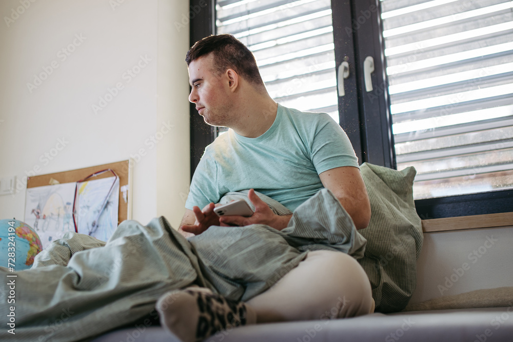 Young man with down syndrome sitting in bed, looking at smartphone in morning. Morning routine for man with disability.