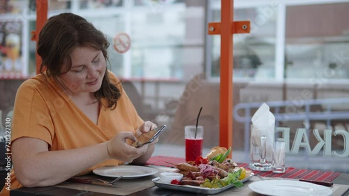 Plus size woman takes photographs of national Balkan dishes on terrace of cafe. Traveler takes pictures of appetizing assortment of grilled meats and vegetables on smartphone from different angles. photo