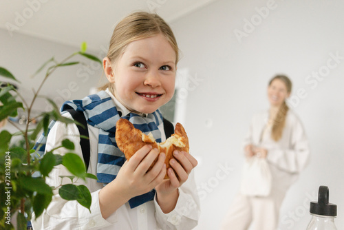 Playful girl eating croissant with mother in background at home photo