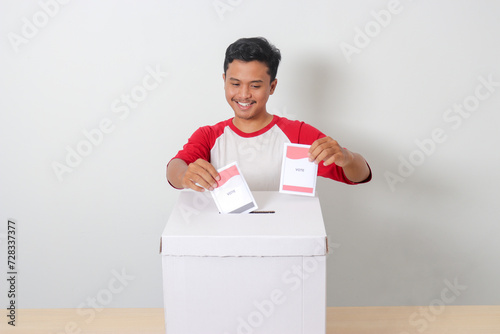 Portrait of excited Asian man inserting and putting the voting paper into the ballot box. General elections or Pemilu for the president and government of Indonesia. Isolated image on white background photo