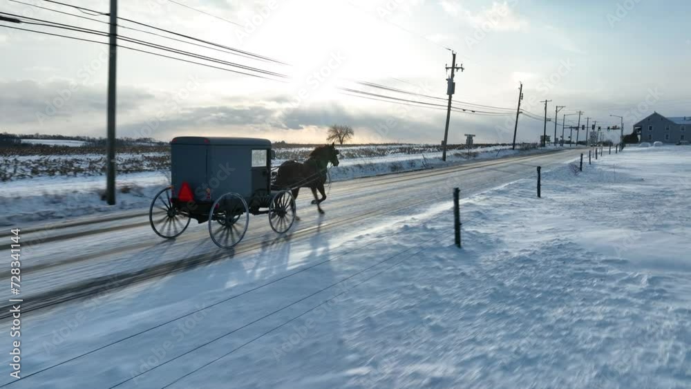 Amish buggy on snowy road in Lancaster County, PA, under a sunset sky. Aerial tracking shot as snow drifts on rural USA road. Amish buggy on snowy road in Lancaster County, PA, under a sunset sky. Aerial tracking shot as snow drifts on rural USA road.