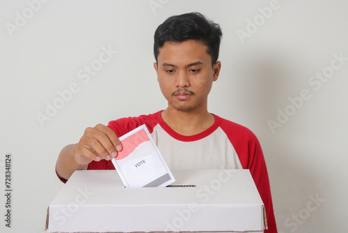 Portrait of excited Asian man inserting and putting the voting paper into the ballot box. General elections or Pemilu for the president and government of Indonesia. Isolated image on white background photo