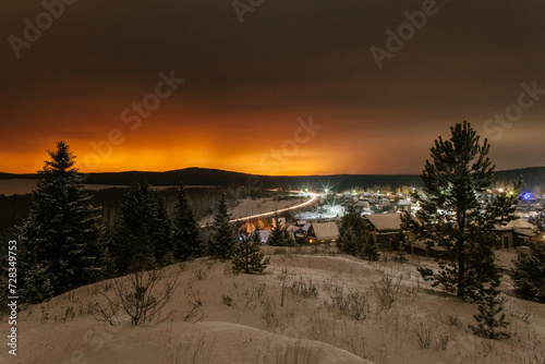 Mountain landscape with village in winter, houses and pine trees covered snow at night. Scenery of ski resort with lights.
