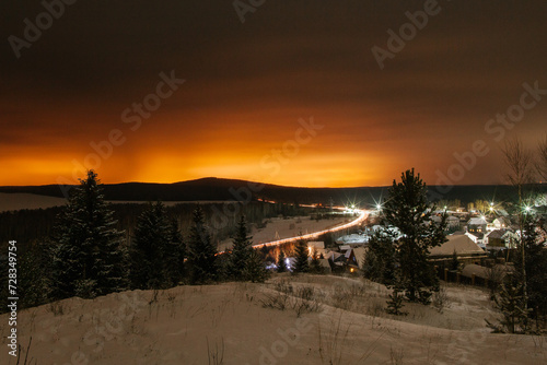Mountain landscape with village in winter, houses and pine trees covered snow at night. Scenery of ski resort with lights.