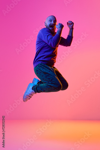 Full-length image of bearded bald man in casual clothes emotionally jumping against pink background in neon light. Concept of human emotions, facial expression, lifestyle