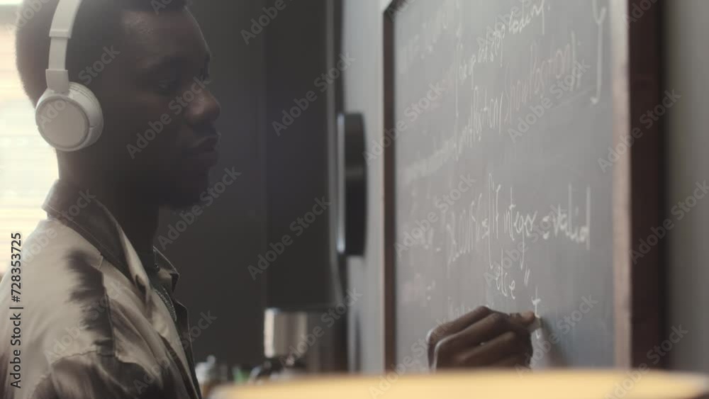 African American IT specialist in wireless headphones writing computer algorithm on chalkboard in office African American IT specialist in wireless headphones writing computer algorithm on chalkboard in office