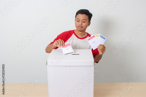 Portrait of excited Asian man inserting and putting the voting paper into the ballot box. General elections or Pemilu for the president and government of Indonesia. Isolated image on white background photo