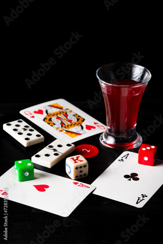 Top view of cards, dice and dominoes with glass of liquor on black background, vertical with copy space