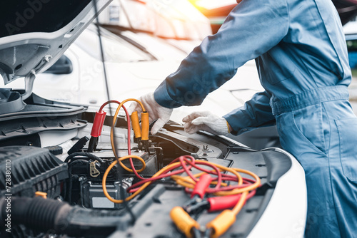 A professional mechanic in a blue uniform jump starts a car with jumper cables in a workshop setting..