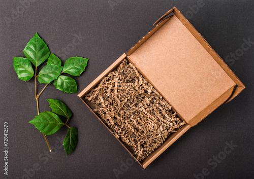 Mockup. Open cardboard box with paper filling on black background with green leaves top view. © Polina Ponomareva
