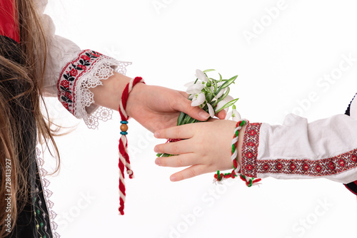Bulgarian kids boy and girl in traditional ethnic folklore costumes with hands with bouquet of spring flowers snowdrops and handcraft wool bracelet martenitsa symbol of Baba Marta March holiday