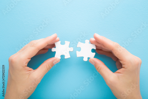 Young adult woman hand fingers holding and connecting different two white puzzle pieces on light blue table background. Pastel color. Closeup. Compatibility concept. Point of view shot. Top down view.