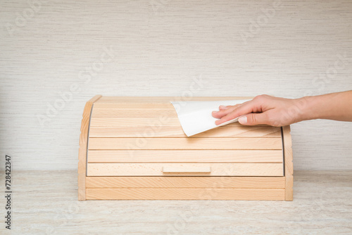Young adult woman hand holding dry white paper napkin and wiping wooden bread box on table top at home kitchen. Closeup. Regular cleanup. Front view.