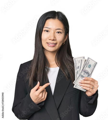 Young Chinese woman with dollars in suit smiling and pointing aside, showing something at blank space.