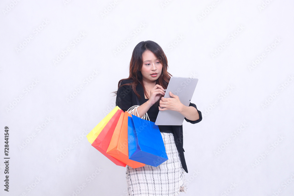 young beautiful woman customer 20s formal serious office holding pen cross checking order on tablet looking ahead and carrying shopping paper bag isolated white background