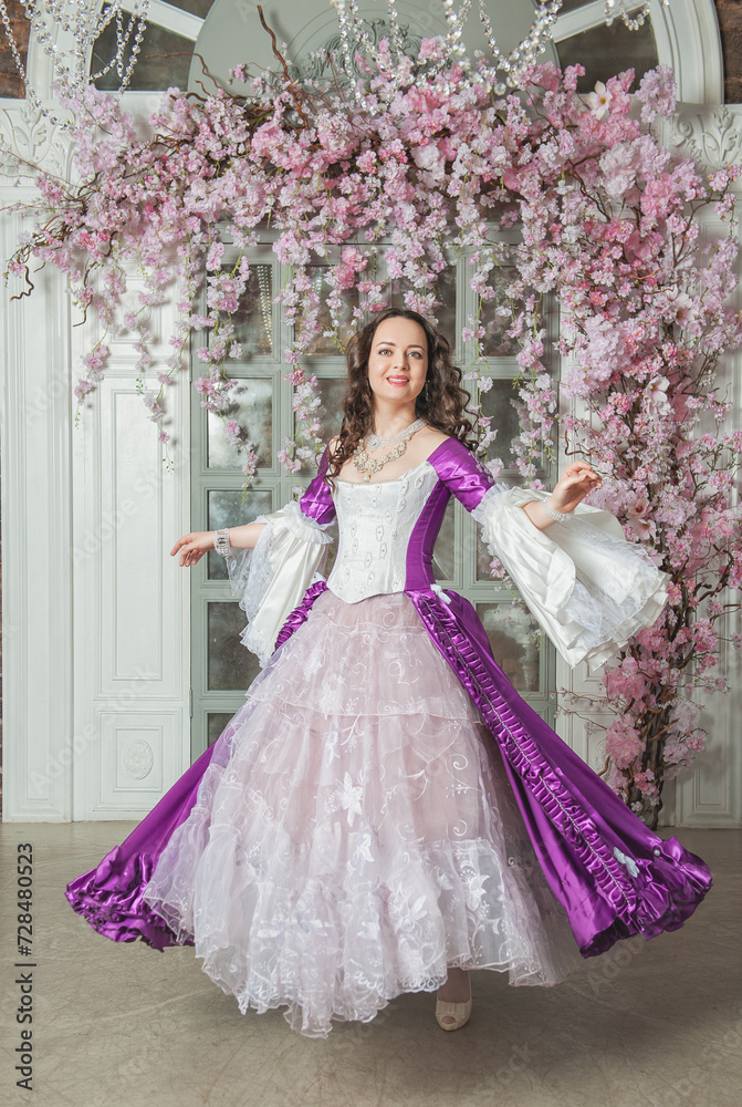 Beautiful woman in fantasy white and purple rococo style medieval dress dancing near door with pink flowers Beautiful woman in fantasy white and purple rococo style medieval dress dancing near door with pink flowers