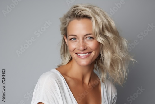 A woman with a bright smile and sun-kissed wavy hair, wearing a simple white top, exuding confidence and natural beauty against a soft grey background