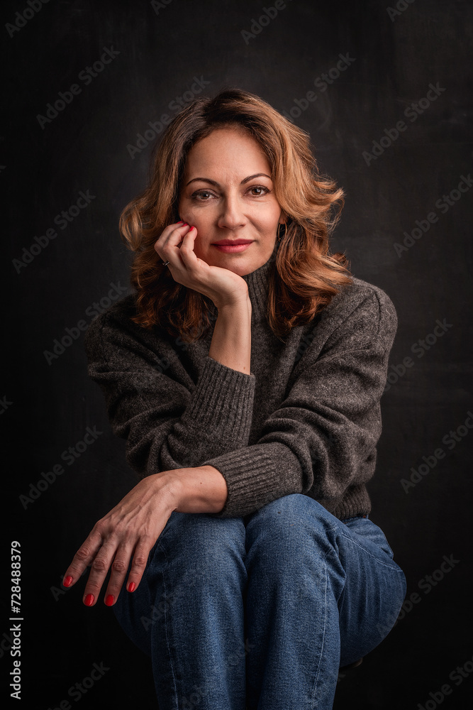 Attractive mid aged woman wearing casual clothes and sitting against dark background Attractive mid aged woman wearing casual clothes and sitting against dark background