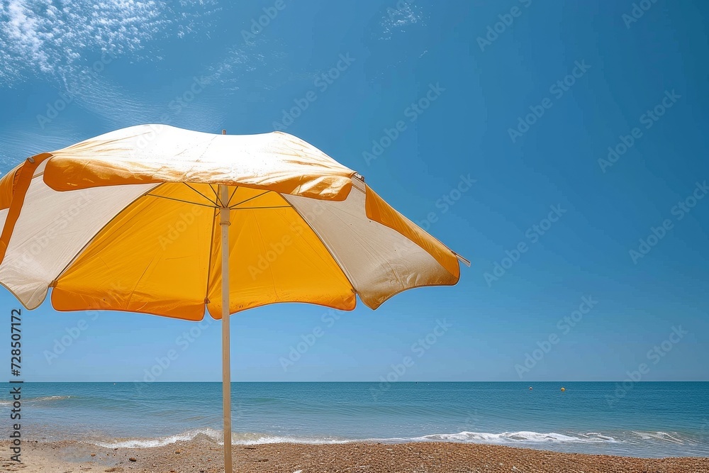 Under the bright blue sky, a vibrant yellow and white umbrella provides shelter on the sandy beach, while the crashing waves of the ocean create a serene soundtrack for a peaceful day in the sun