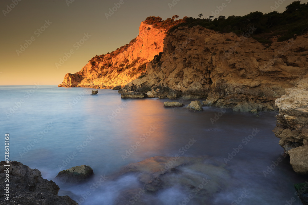 Golden sunlight illuminates a rugged cliff face above a serene Mediterranean cove with silky blue waters Golden sunlight illuminates a rugged cliff face above a serene Mediterranean cove with silky blue waters