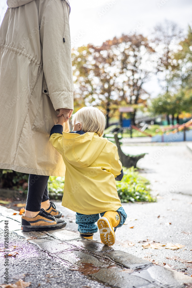 Small bond infant boy wearing yellow rubber boots and yellow waterproof raincoat walking in puddles on a overcast rainy day holding her mother's hand. Mom with small child in rain outdoors Small bond infant boy wearing yellow rubber boots and yellow waterproof raincoat walking in puddles on a overcast rainy day holding her mother's hand. Mom with small child in rain outdoors