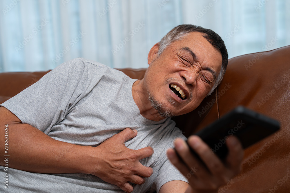 Senior male asian presses hand to his chest has a heart attack suffers from unbearable pain in the living room, lonely men try using the smartphone to call for Emergency help.