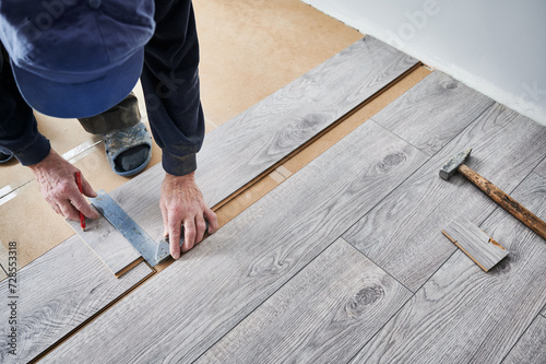 Man preparing laminate plank for floor installation in apartment under renovation. Close up of male construction worker using metal ruler and pencil while drawing line on laminate flooring board.