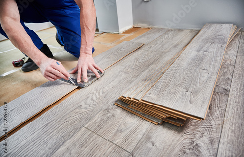 Man preparing laminate plank for floor installation in apartment under renovation. Close up of male worker using metal construction ruler and pen while drawing line on laminate flooring board.