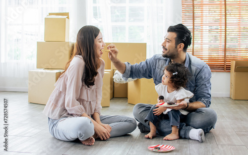 Three people, father, mother, little daughter moving to new house, sitting on floor, eating cookies together with happiness with background of cardboards, unopened boxes. Family, Real estate concept. photo