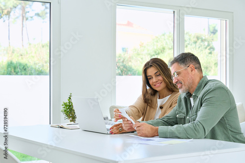 Happy middle aged couple mature man and woman calculating investments and financial expenses, money savings, paying bills online, using laptop computer sitting at home table in living room. Copy space