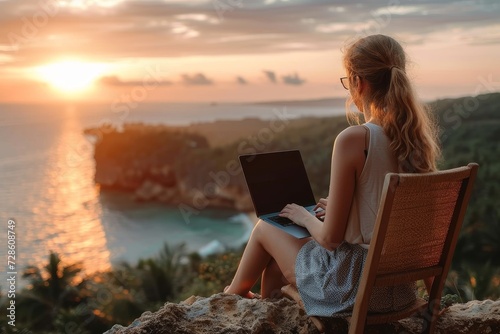 A girl embraces the serenity of nature as she sits on a rock, typing away on her laptop against the backdrop of a stunning sunset over the ocean