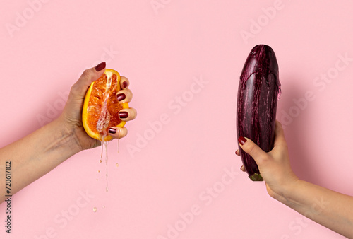 Female hand with half of juicy orange and eggplant on pink background. Sex concept.