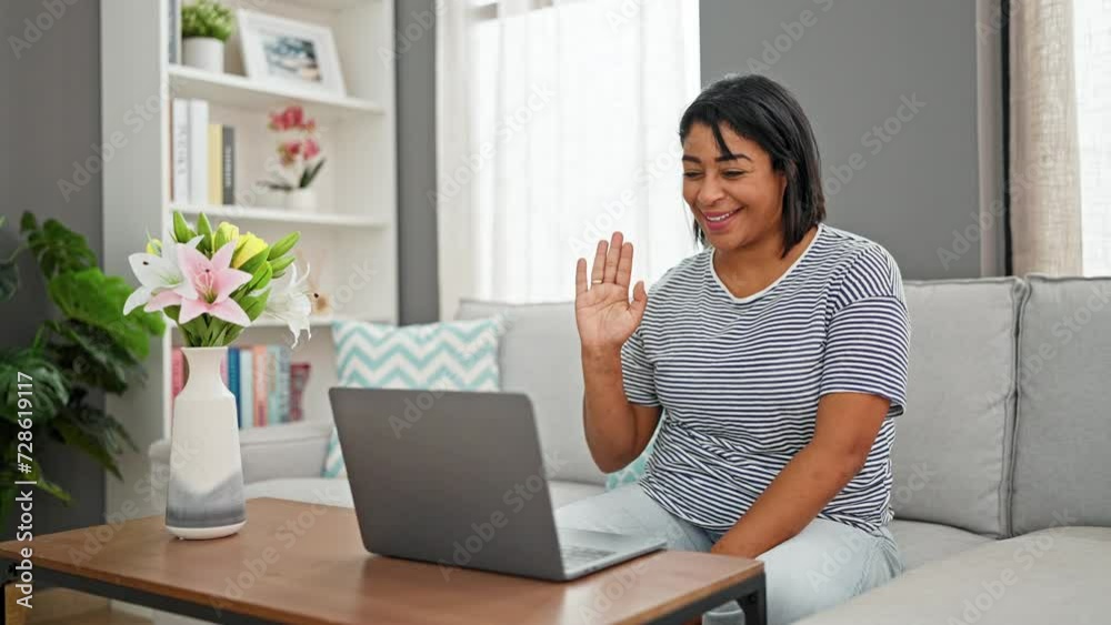 Smiling woman sitting on sofa waving at laptop camera in a cozy living room setup Smiling woman sitting on sofa waving at laptop camera in a cozy living room setup