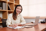 Beaming Asian office worker shares a moment of success, her smile reflecting a job well done at laptop. Success lights up face of professional as she glances up from laptop in well-appointed office