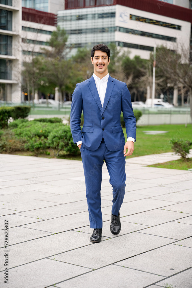 A full-length shot of a cheerful Hispanic businessman walking confidently in a blue suit, with a bustling cityscape providing a dynamic backdrop to his active lifestyle.
