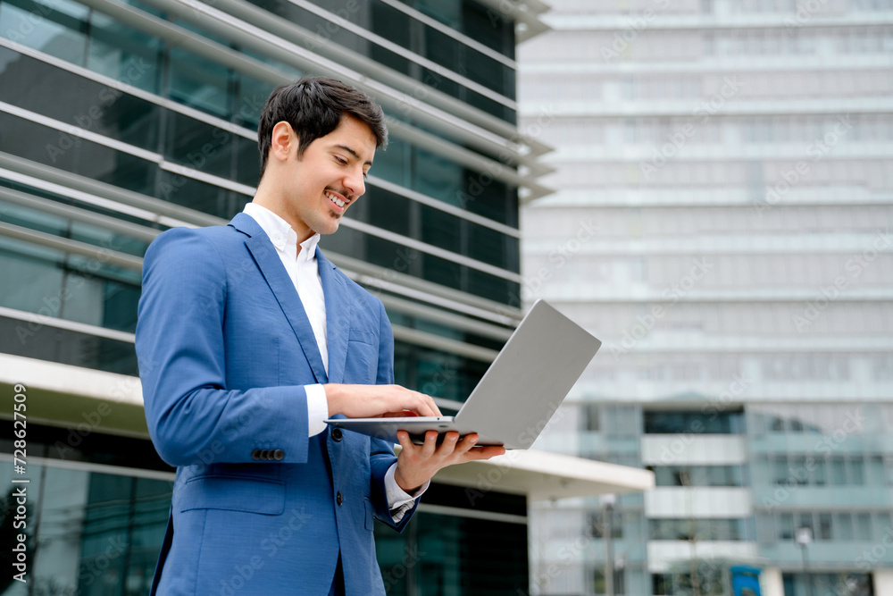 Fototapeta premium A smiling Hispanic businessman stands with an open laptop in front of a modern office, embodying the concept of accessible technology and business efficiency in today's fast-paced world.