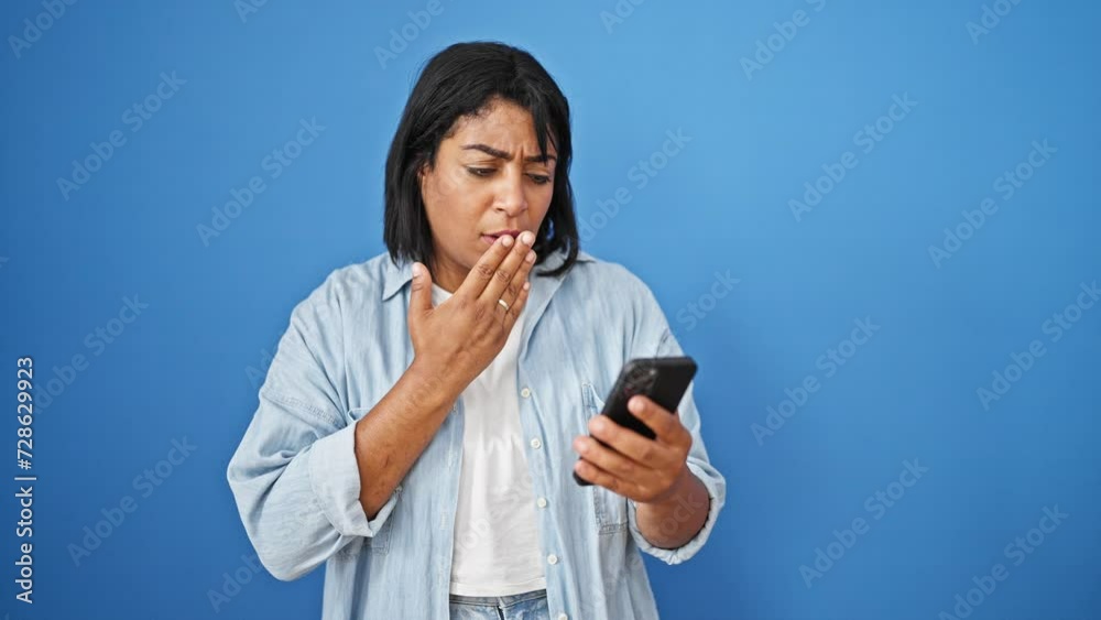 Shocked hispanic woman looking at smartphone against blue background Shocked hispanic woman looking at smartphone against blue background