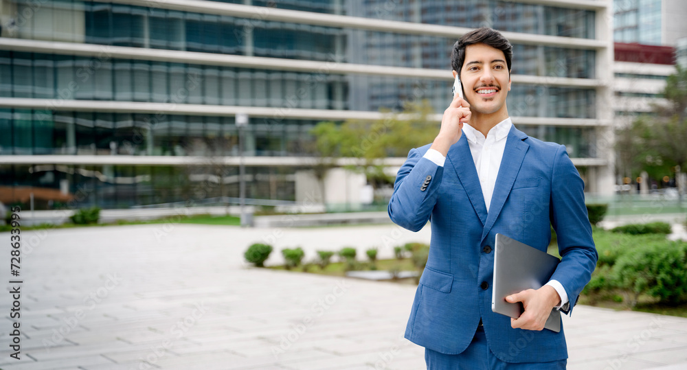 Smiling Hispanic businessman in a blue suit makes a call on his smartphone while holding a laptop, portraying a successful, connected, and mobile entrepreneur in an urban setting. Multitasking concept Smiling Hispanic businessman in a blue suit makes a call on his smartphone while holding a laptop, portraying a successful, connected, and mobile entrepreneur in an urban setting. Multitasking concept
