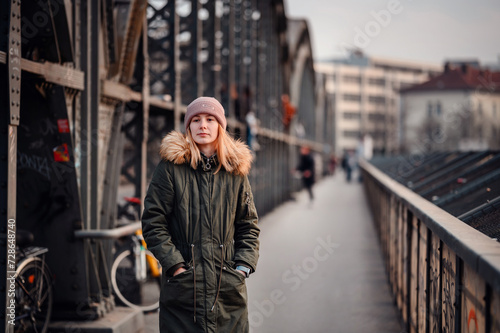 A young person in winter attire leans on a city bridge railing, gazing thoughtfully