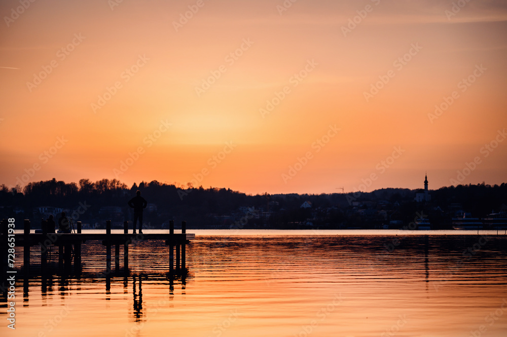 A person stands on a dock, enjoying the vibrant orange hues of sunset reflecting on a peaceful lake