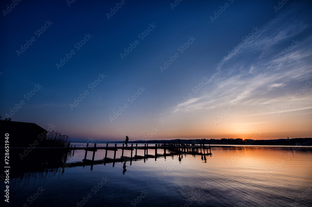 Silhouette of a person on a pier against the twilight sky reflected on a calm lake's surface