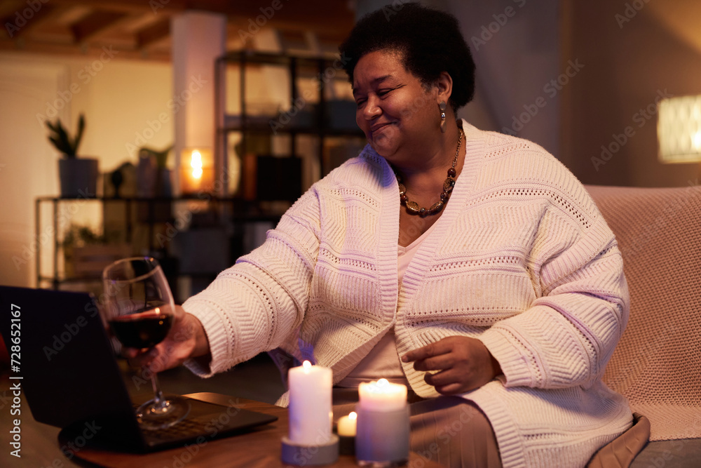 Side view portrait of mature Black woman clinking wine glass to screen while talking by video chat and enjoying relaxing evening at home copy space